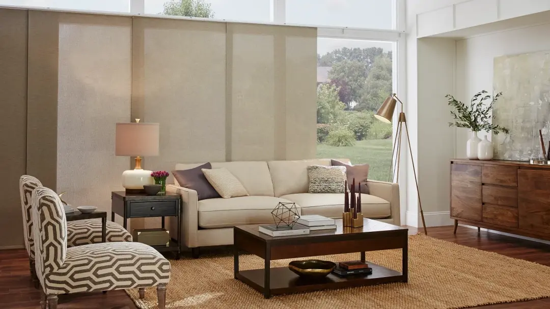 Light beige Alta Window Fashions sliding panel track shades covering a wall of windows in a bright living room with a beige sofa, patterned accent chairs and wood furniture